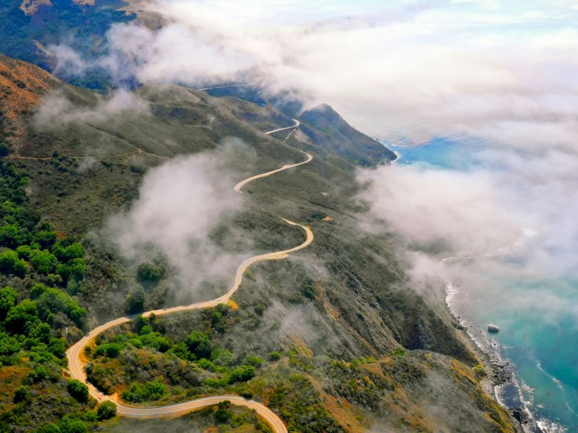 A winding cliffside road snakes along a lush green mountainside, vanishing into misty clouds above the blue ocean, creating a dramatic coastal switchback.