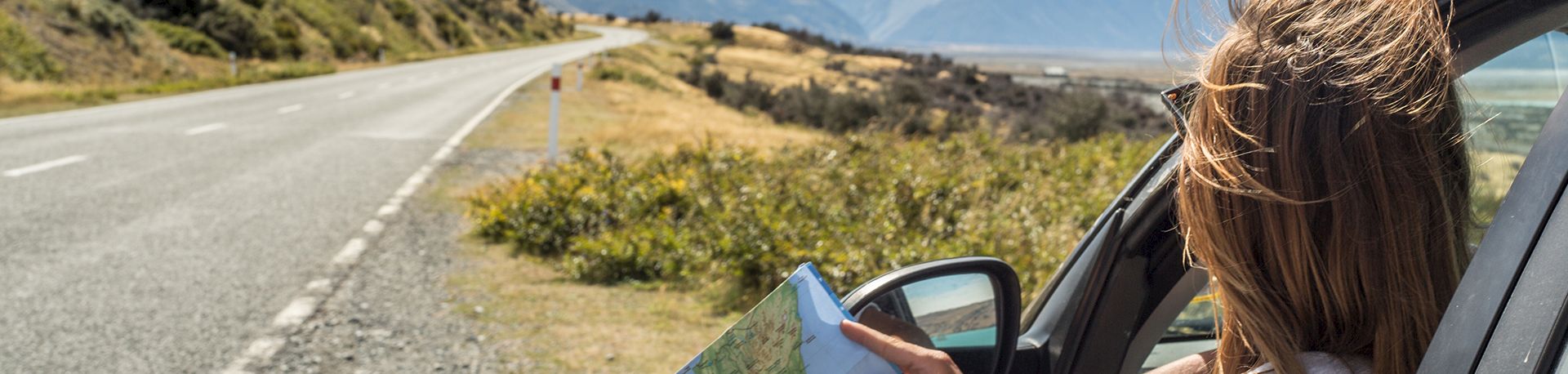 A person seated in a car on a rural highway, looking at a map with mountains in the distance under a sunny sky.