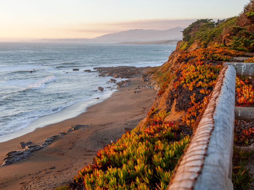 Coastline view with a sandy beach, rocky shore, ocean waves, and a wooden railing along a cliff path at sunset.