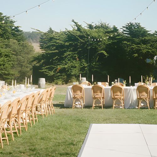 A long rectangular table setup with white linens and many wooden chairs outdoors, string lights overhead, on a grassy area near trees, ready for an event.