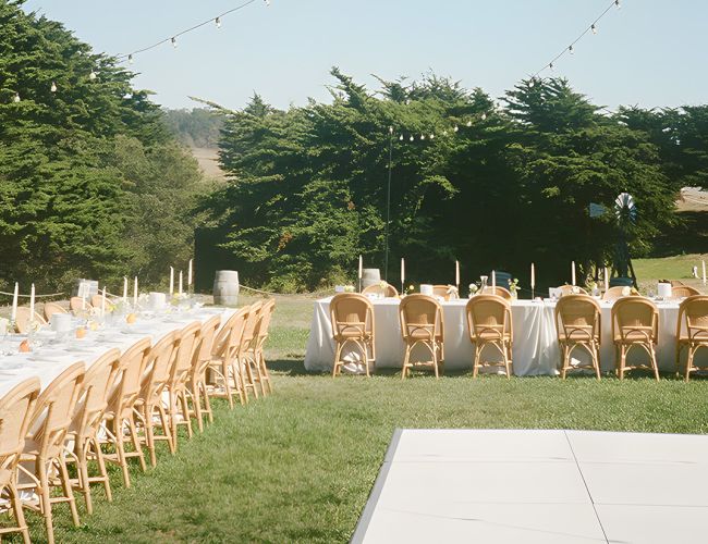 A long rectangular table setup with white linens and many wooden chairs outdoors, string lights overhead, on a grassy area near trees, ready for an event.