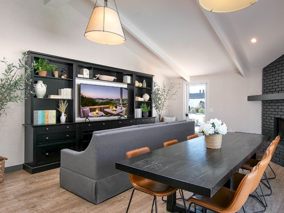 A modern open‑plan living and dining area with a dark shelving unit, a gray sectional sofa, a long dining table, and warm pendant lighting.