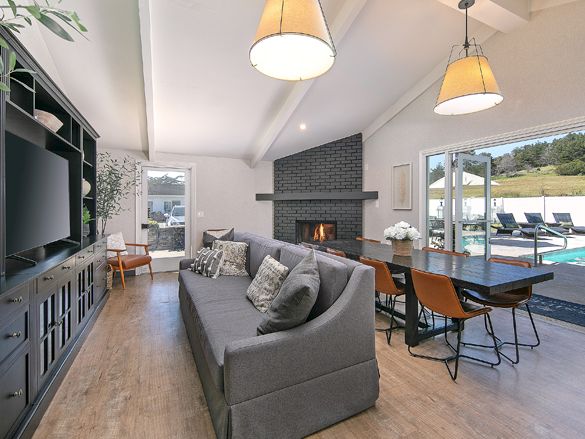 Cozy modern living room with a gray sofa, built-in shelving, fireplace, TV unit, and a bright dining area near large glass doors opening to a deck.