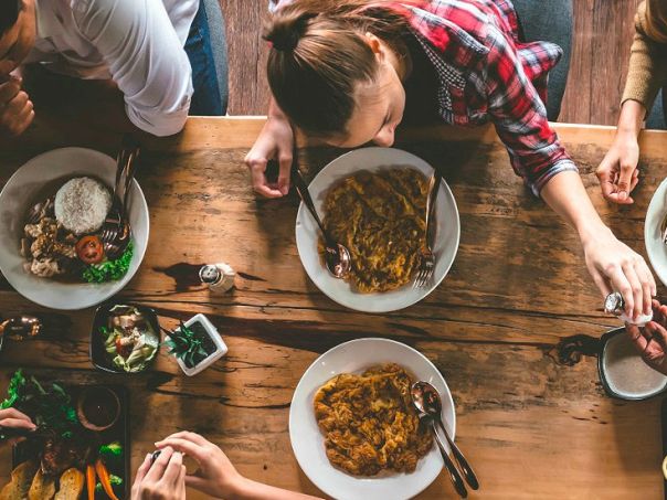 People sit around a wooden table sharing plates of food, including curry and sides, as they reach for bites and chat together.