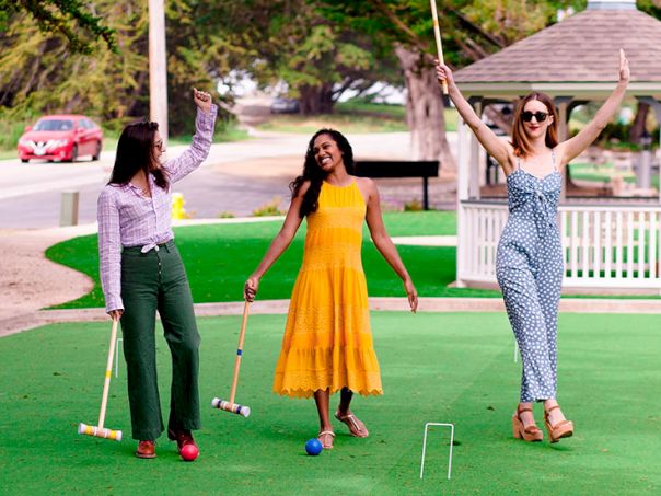 Three women play mini-golf on a sunny day; one waves a broom, another holds a club, and the third arms raised in celebration.