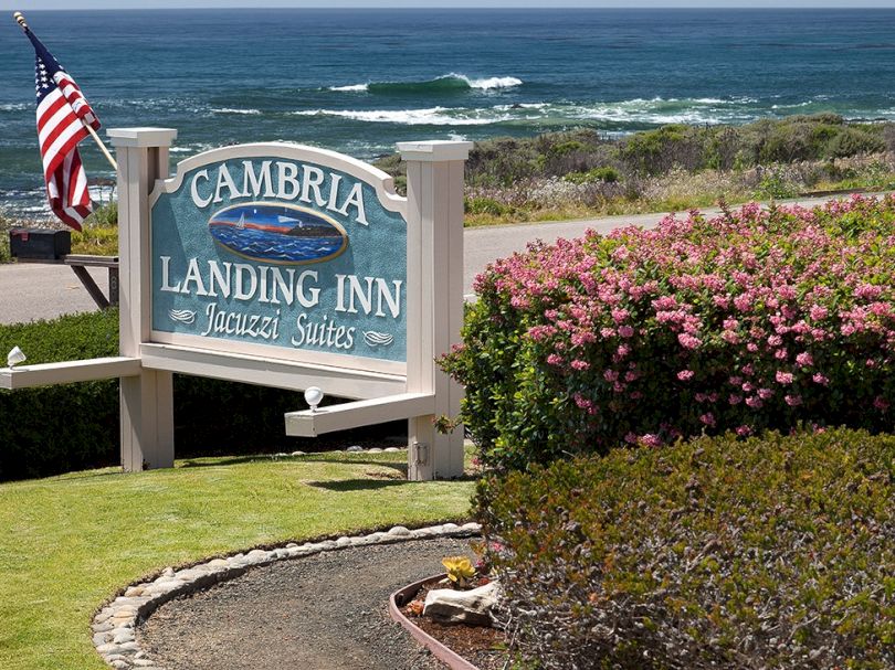 A seaside inn sign reads &ldquo;Cambria Landing Inn&rdquo; near a coastal garden with waves in the background.