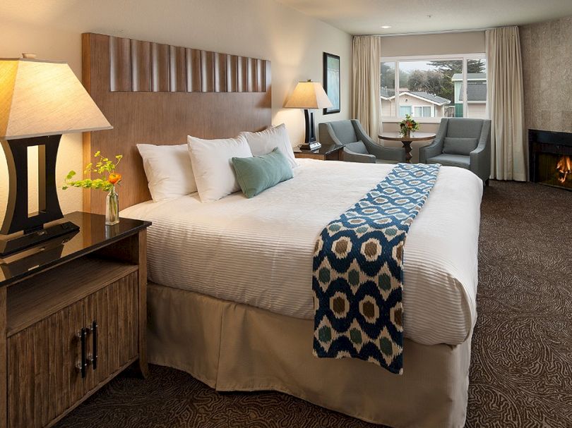 A cozy hotel room with a king bed, geometric throw, two nightstands with lamps, a windowed seating area, and warm, neutral decor.