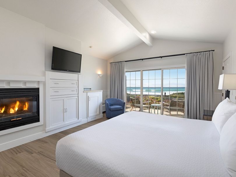 Cozy hotel room with a lit fireplace, white decor, a flat-screen TV, built-in storage, a chair, and a balcony offering ocean views.