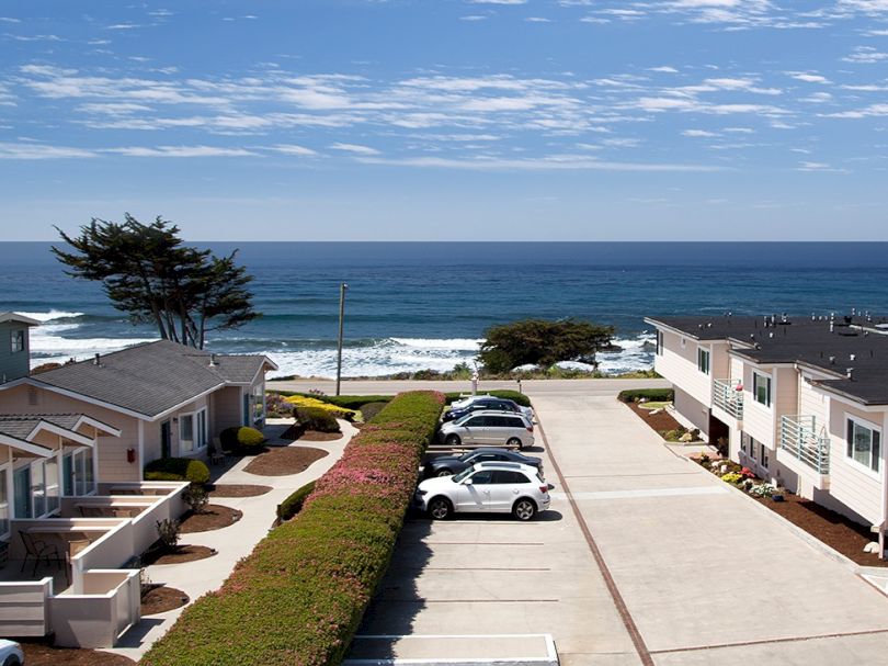 Coastal neighborhood with row houses along a parking lot, grassy hedges, and a view of the ocean under a blue sky, waves near the shore.