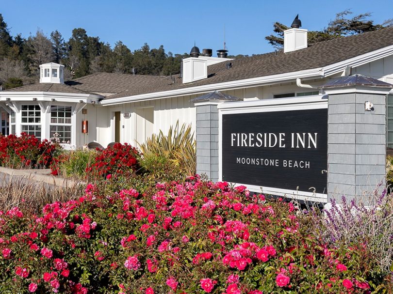 A coastal motel with a sign reading &ldquo;Fireside Inn&rdquo; by Bright pink flowers and shrubs, white buildings, and a clear blue sky, on Moonstone Beach.
