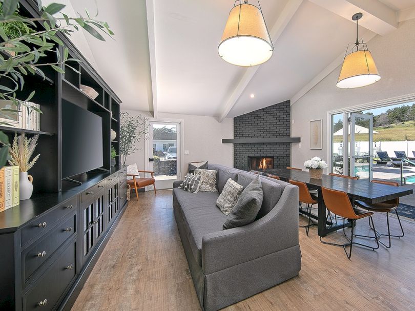 Modern open-concept living room with a gray sofa, built-in media wall, and a cozy dining nook by a brick fireplace, overlooking outdoors.