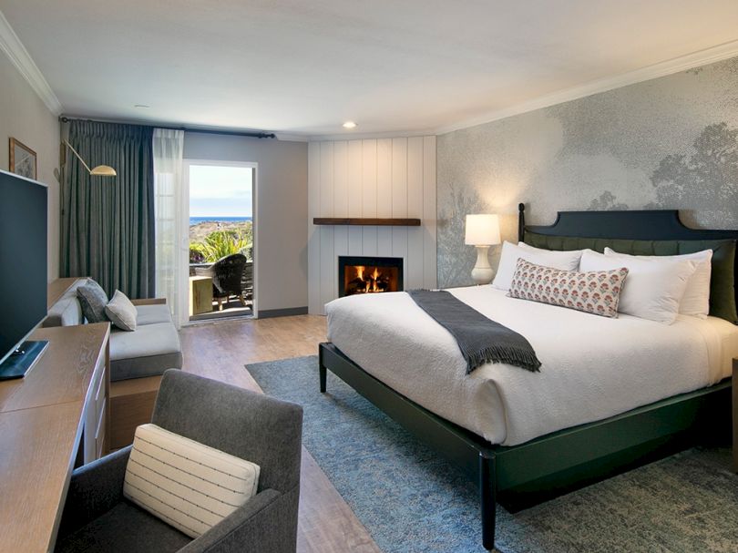 A modern hotel room with a king bed, fireplace, seating area, and balcony view, calm blue-gray decor, and soft lighting.