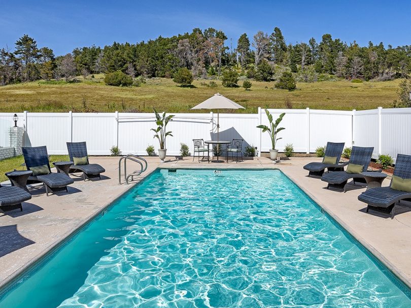 Swimming pool surrounded by lounge chairs, blue water sparkling under a sunny sky, with a white fence and green hills in the background. end sentence.