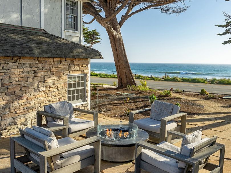 A cozy outdoor seating area with stone walls, four cushioned chairs around a round fire pit, overlooking a sunny beach and ocean.