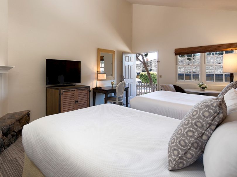 A bright hotel room with two queen beds, a desk and chair, a small dresser with a TV, and a balcony with a view outside.