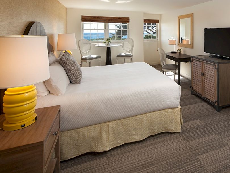 A hotel room with a large bed, yellow lamp, bedside tables, a desk area, a sofa, a flat-screen TV, and a windowed view by the ocean, relaxing vibe.
