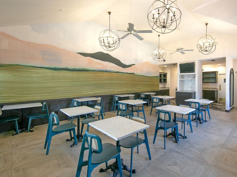 A bright, modern cafe with pastel-blue chairs, white-tleck tables, a coastal mural, and geometric pendant lights across an airy dining area.