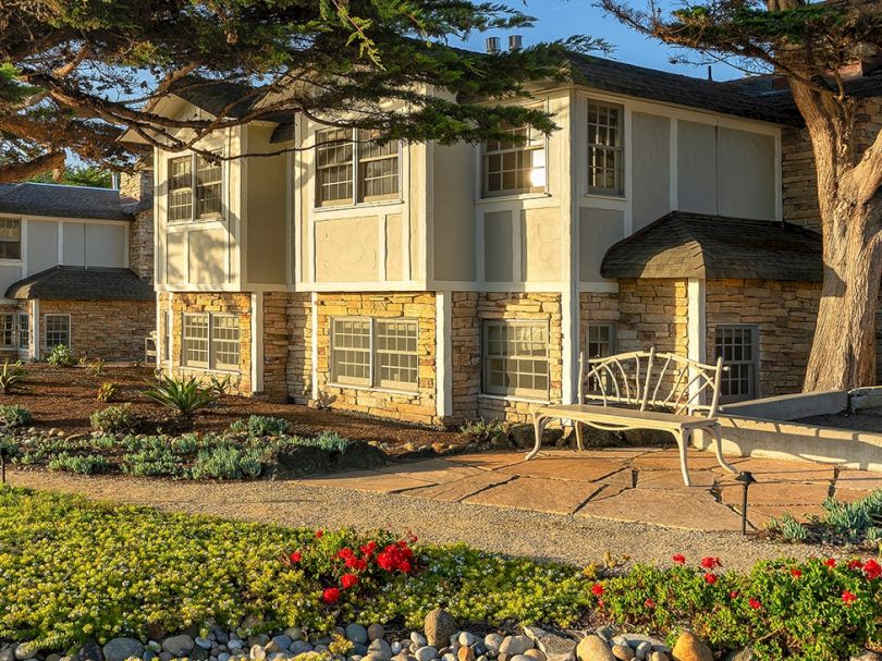 A two-story beige house with large windows, a low stone foundation, a metal ramp, and landscaped front yard with flowers and a tree.