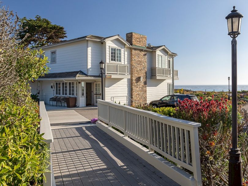 A seaside house with a wooden ramp, white siding, stone chimney, and a bright blue sky by the coast, near a lamppost and garden. Channel: 140 chars max.