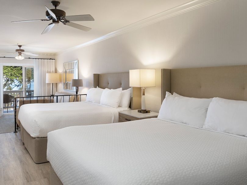 A bright hotel room with two large beds, nightstands with lamps, a beige headboard, ceiling fans, and a balcony view in the background.