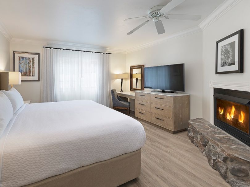 A bright hotel-style bedroom with a king bed, dresser, flat-screen, fireplace, stone accent, and neutral decor, cozy and modern.