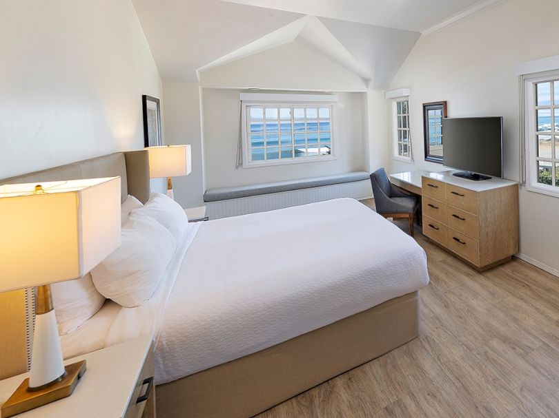 A bright hotel room with a queen bed, two nightstands, a desk with a chair, a dresser with a TV, and a window-lit seating area, stylish and cozy.