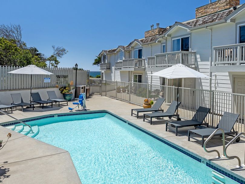 A bright outdoor pool area with clear water, lounge chairs, umbrellas, and a row of light apartment buildings in the background, sunny day.