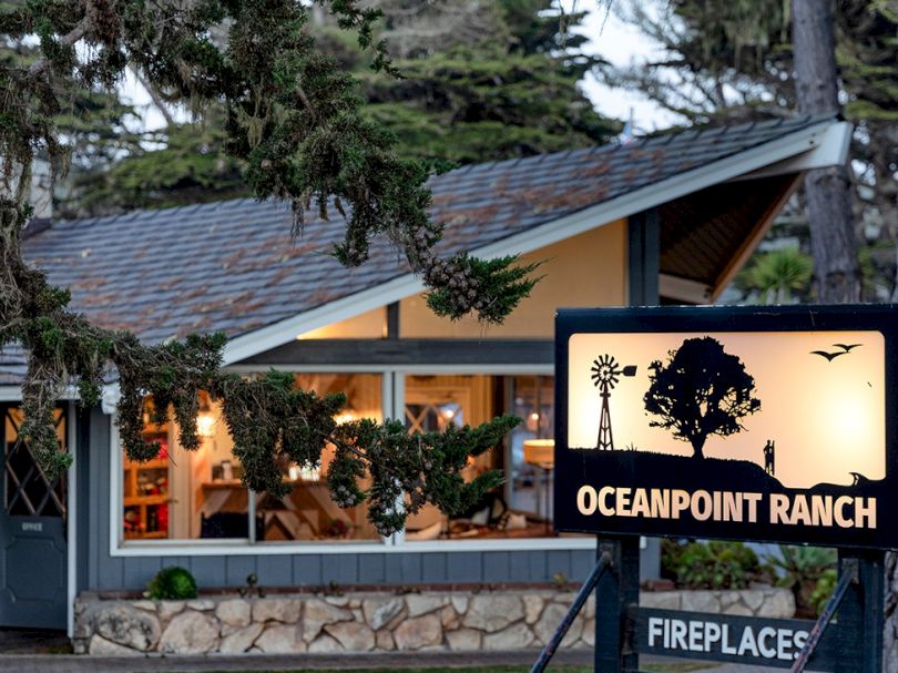 A cozy ranch-style cafe with warm lights behind a stone wall, nestled by trees; sign reads Oceanpoint Ranch with a fireside/beach vibe.