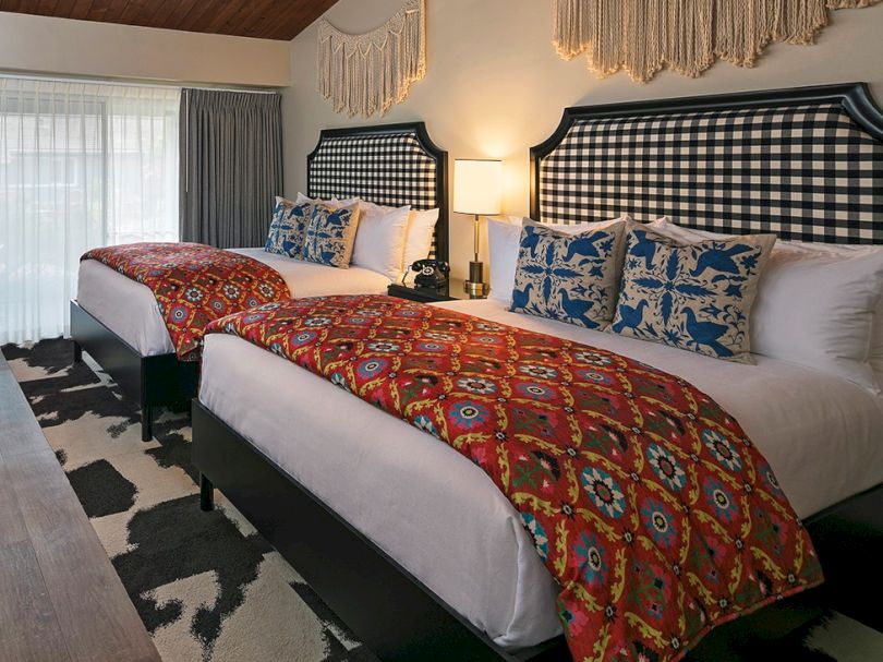 Two comfy twin beds with black-and-white check headboards, red patterned runners, blue throw pillows, a nightstand lamp, and a woven wall hanging in a bright hotel room.