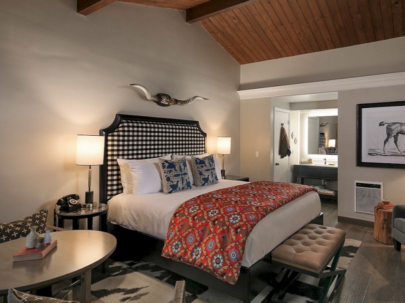 A luxurious hotel room with a king bed, patterned headboard, two lamps, decorative pillows, and a red-orange throw; a seating area and writing desk nearby.