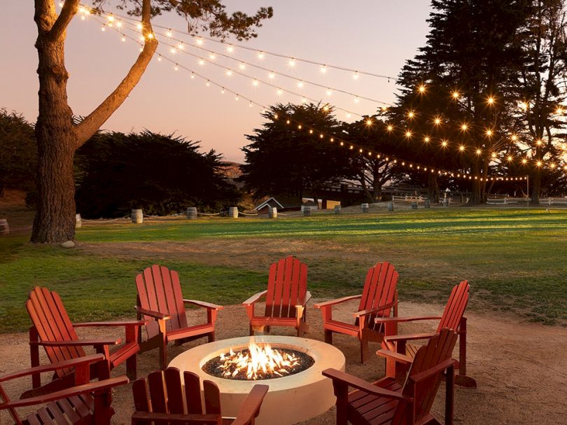 A cozy outdoor fire pit surrounded by red Adirondack chairs under string lights at dusk in a park.