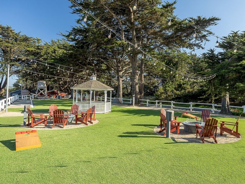 A sunny outdoor garden with a gazebo, Adirondack chairs, string lights, and a white fence around lush green grass and tall trees.
