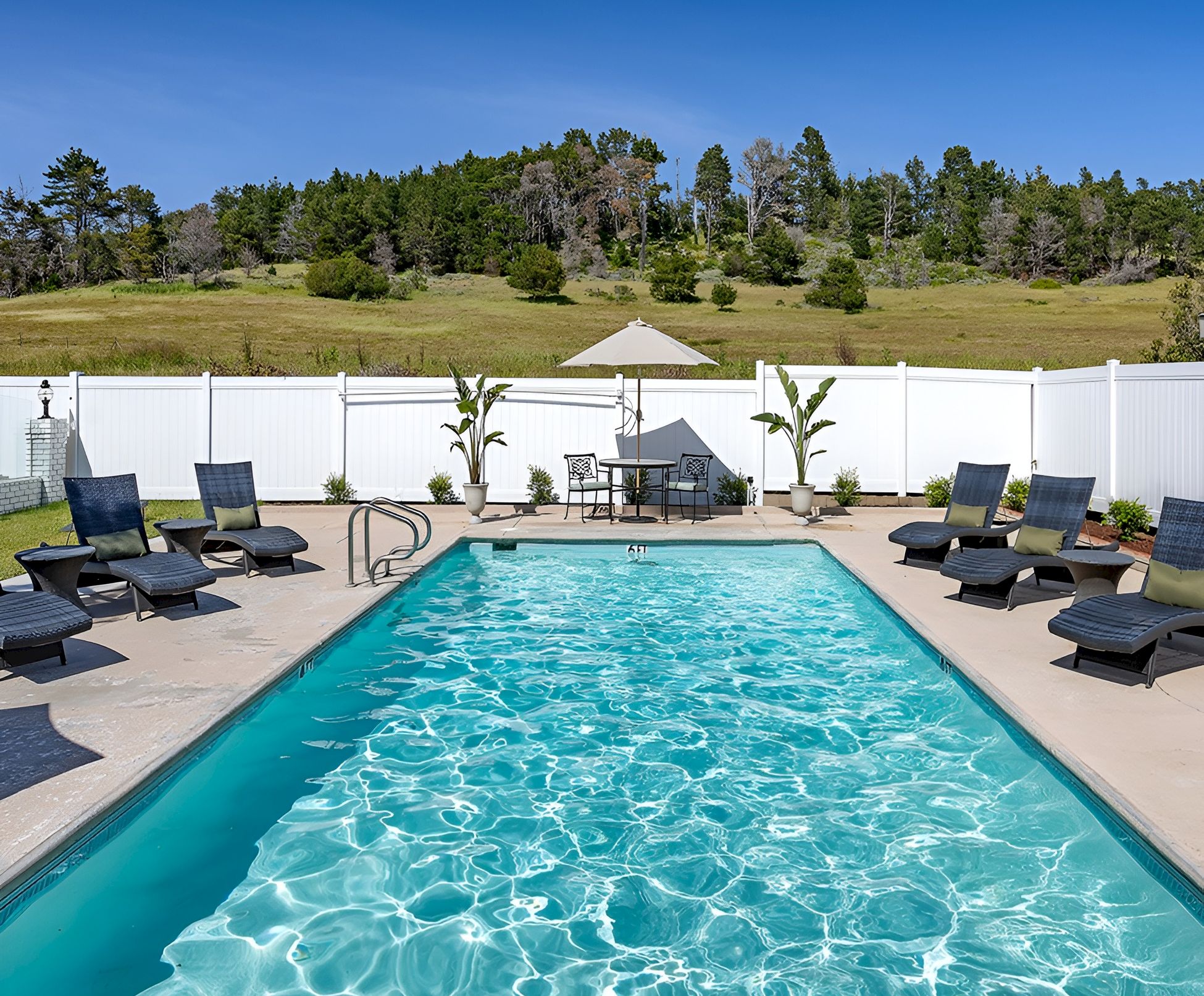 A sunny backyard with a rectangular pool, blue water, black lounge chairs around, a small seating area with an umbrella, and a white fence with a grassy hill in the background.