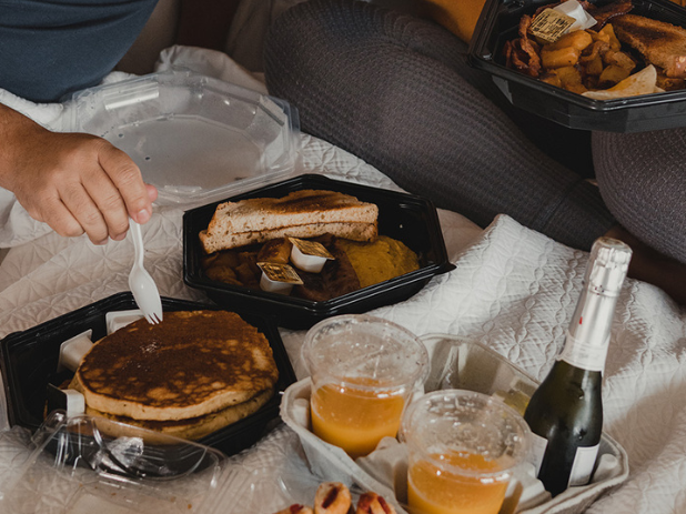 Two people eating in bed with takeout trays: pancakes, sides, orange juice, and a bottle of sparkling wine on a blanket picnic. The scene looks cozy and casual.