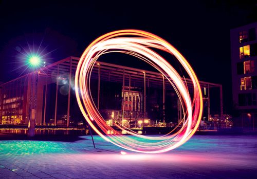 A long-exposure shot of a person spinning a light orb, creating a glowing circular arc of rainbow colors over a city plaza at night.