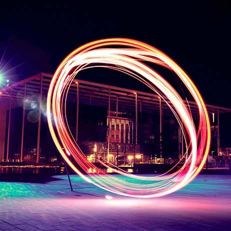 A long-exposure shot of a person spinning a light orb, creating a glowing circular arc of rainbow colors over a city plaza at night.