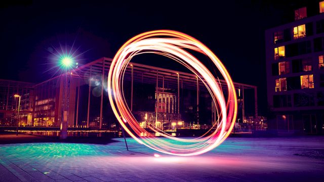 A long-exposure shot of a person spinning a light orb, creating a glowing circular arc of rainbow colors over a city plaza at night.