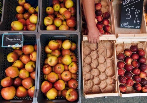 A market stall with apples in crates and boxes of nectarines and plums; a hand reaches toward the produce, a chalkboard sign visible.