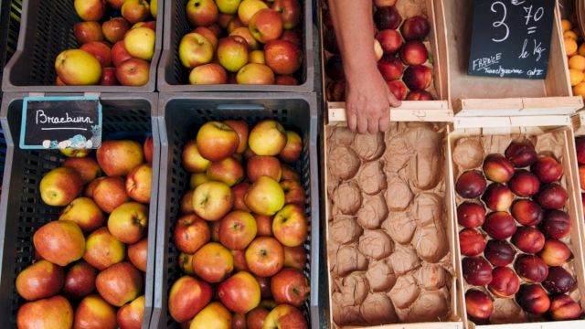 A market stall with apples in crates and boxes of nectarines and plums; a hand reaches toward the produce, a chalkboard sign visible.