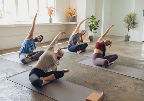 People doing a yoga class in a bright studio, various seated side stretches on mats with blocks nearby.