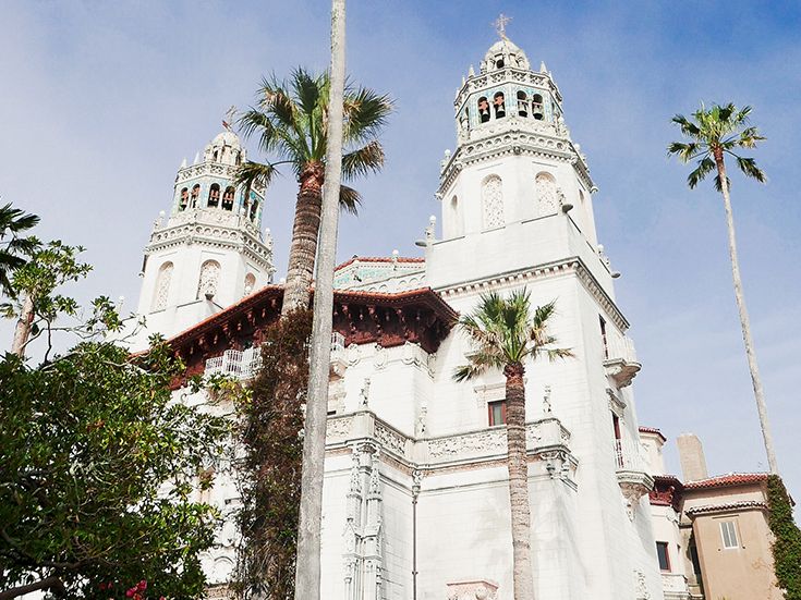A white mission-style building with tall towers, palm trees nearby, and a clear blue sky in the background.