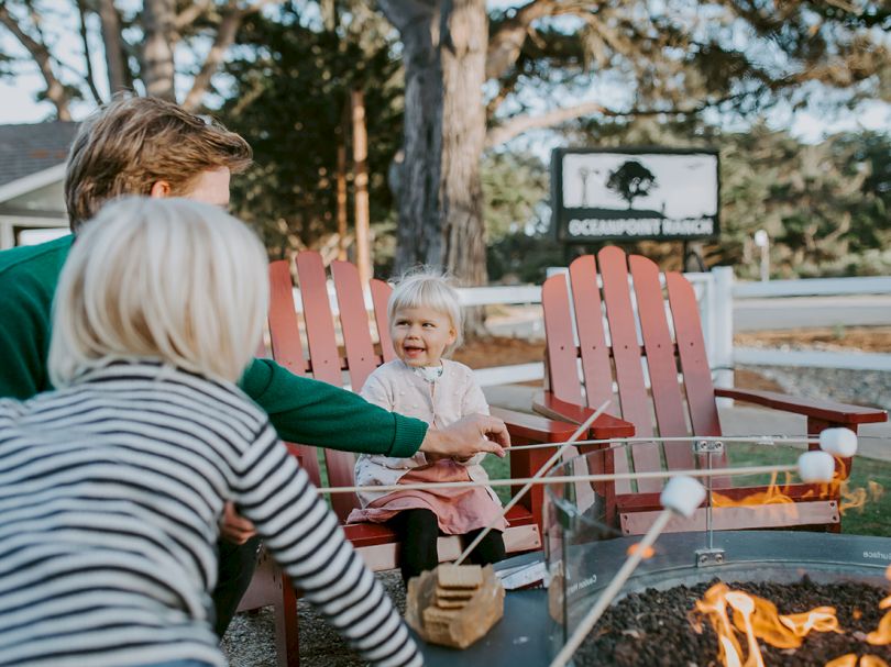 Two adults and a toddler sit around a campfire outdoor, smiling as the child sits in a red chair, roasting marshmallows.