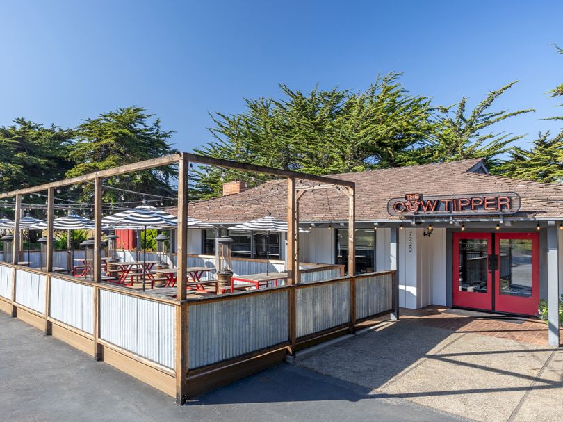 A small outdoor-seating restaurant or cafe with a wooden fenced patio, striped umbrellas, and a red double-door entrance under a simple signage. End.