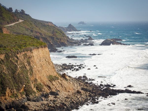 A rugged coastal scene with cliffs, rocky shoreline, and waves crashing against rocks along the blue ocean, under a clear sky.