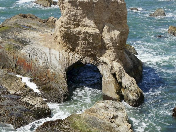 A jagged sea stack rises from turquoise waters along a rocky coastline, with a cave at its base and waves crashing nearby, basked in sunlight.
