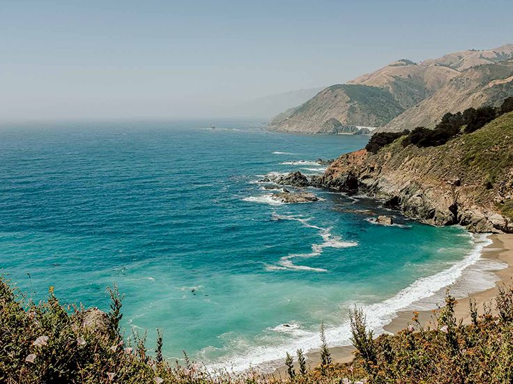 A rugged coastline meeting a turquoise sea, with waves lapping a secluded sandy cove and rocky cliffs fading into distant hazy mountains, vibe of a pristine beach.