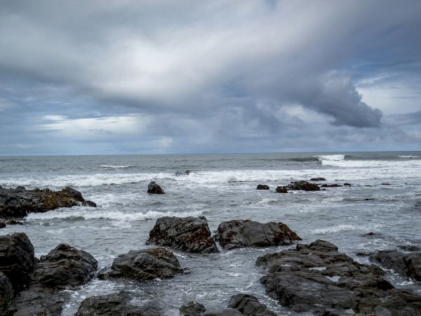 A rocky shoreline with dark, jagged rocks along the sea, waves crashing, and a cloudy, overcast sky above, hinting at approaching rain.