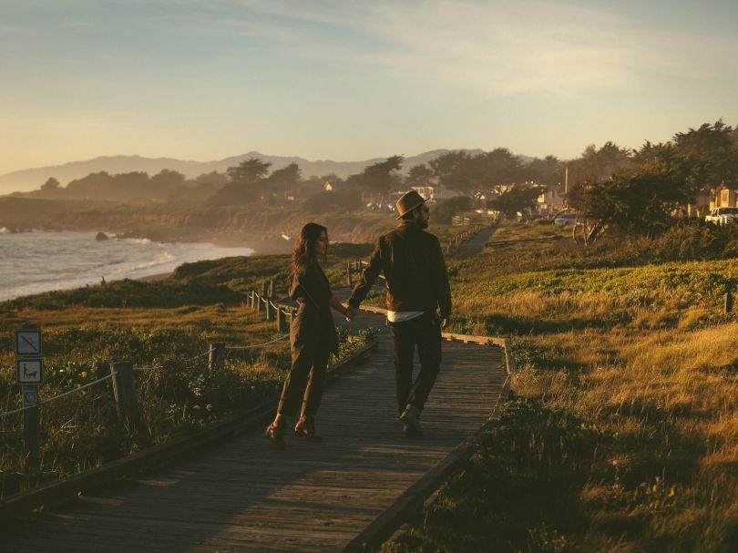 Two people walk a wooden path along a scenic coastline at sunset, waves to the left and grassy cliffs to the right, peaceful and romantic.