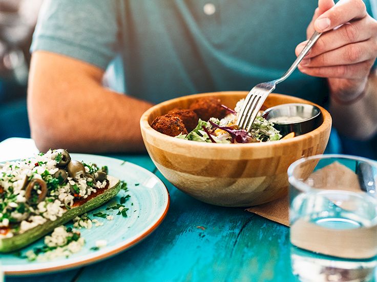 A person eats a wooden bowl meal with a fork while a plate of greens and quinoa sits nearby, and a glass of water rests on a blue table.