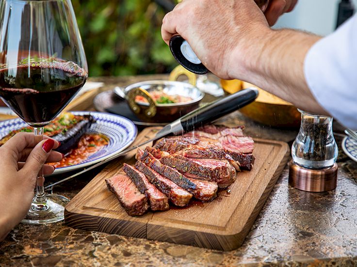 Juicy sliced steak on a wooden board, wine glass in foreground, pepper grinder, and hands seasoning and pouring sauce at a sunny outdoor table.